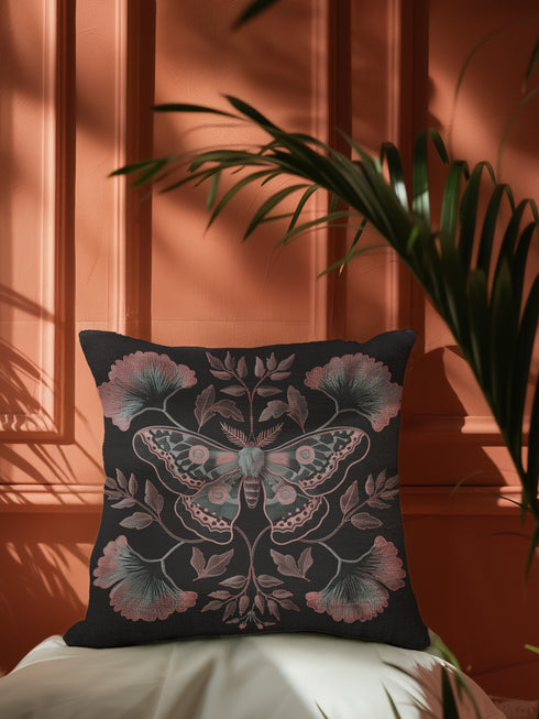 A square decorative pillow featuring an intricately detailed Luna moth in faded rose pink, slate blue and dusty terracotta colors on a black background. The moth is centered in a symmetrical arrangement with ginkgo-inspired leaves and botanical elements surrounding it.